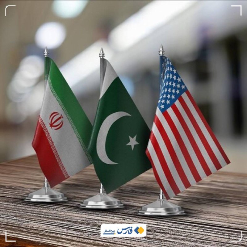 Three small desk flags representing Iran, Pakistan, and the United States stand in a row on a wooden surface against a blurred indoor background.