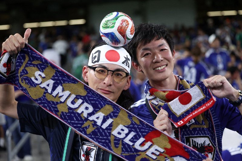 Two men pose smiling at a stadium, one wearing a Japan headband balancing a soccer ball on his head, while displaying a "Samurai Blue" scarf and a Japan pennant.