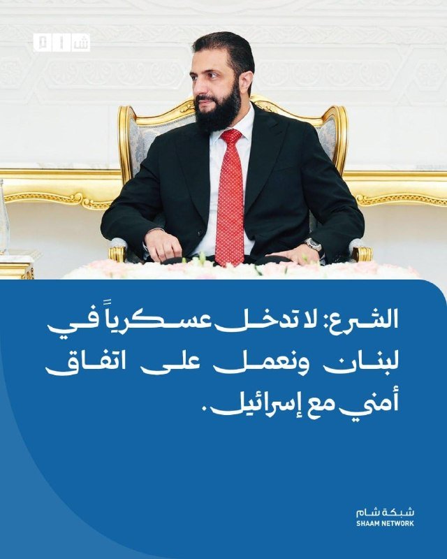 A bearded man in a dark suit and red tie sits in an ornate chair above a blue banner containing Arabic text and a "Shaam Network" logo.