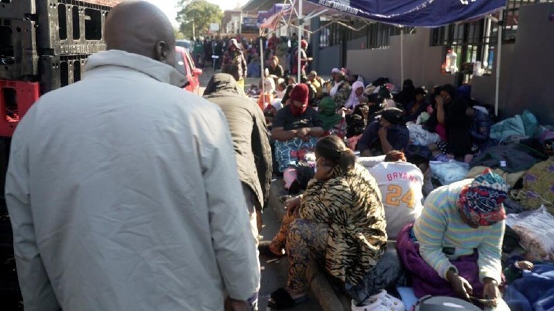 Numerous people sit along a sidewalk in front of a building, surrounded by bundles and belongings, while a man in a light shirt stands in the foreground.