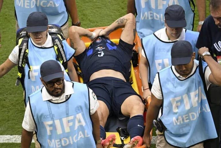 An injured soccer player wearing a dark uniform lies on a stretcher, surrounded by FIFA Medical staff carrying him off the grass field.