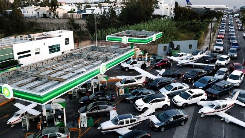 An aerial view shows a BP gas station where multiple cars and small airplanes are parked closely together among the fuel pumps and canopy.
