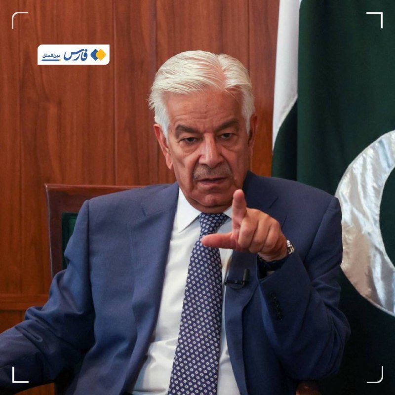 A white-haired man in a blue suit and patterned tie points forward while seated, with a green and white flag visible behind him.