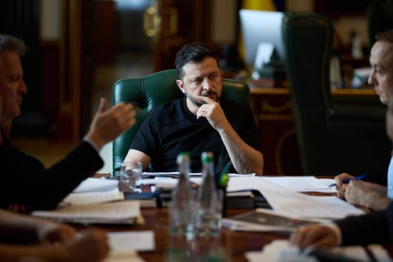 A bearded man in a black shirt sits at a conference table listening to another man gesturing while speaking, with papers, bottles, and water glasses scattered across the surface.