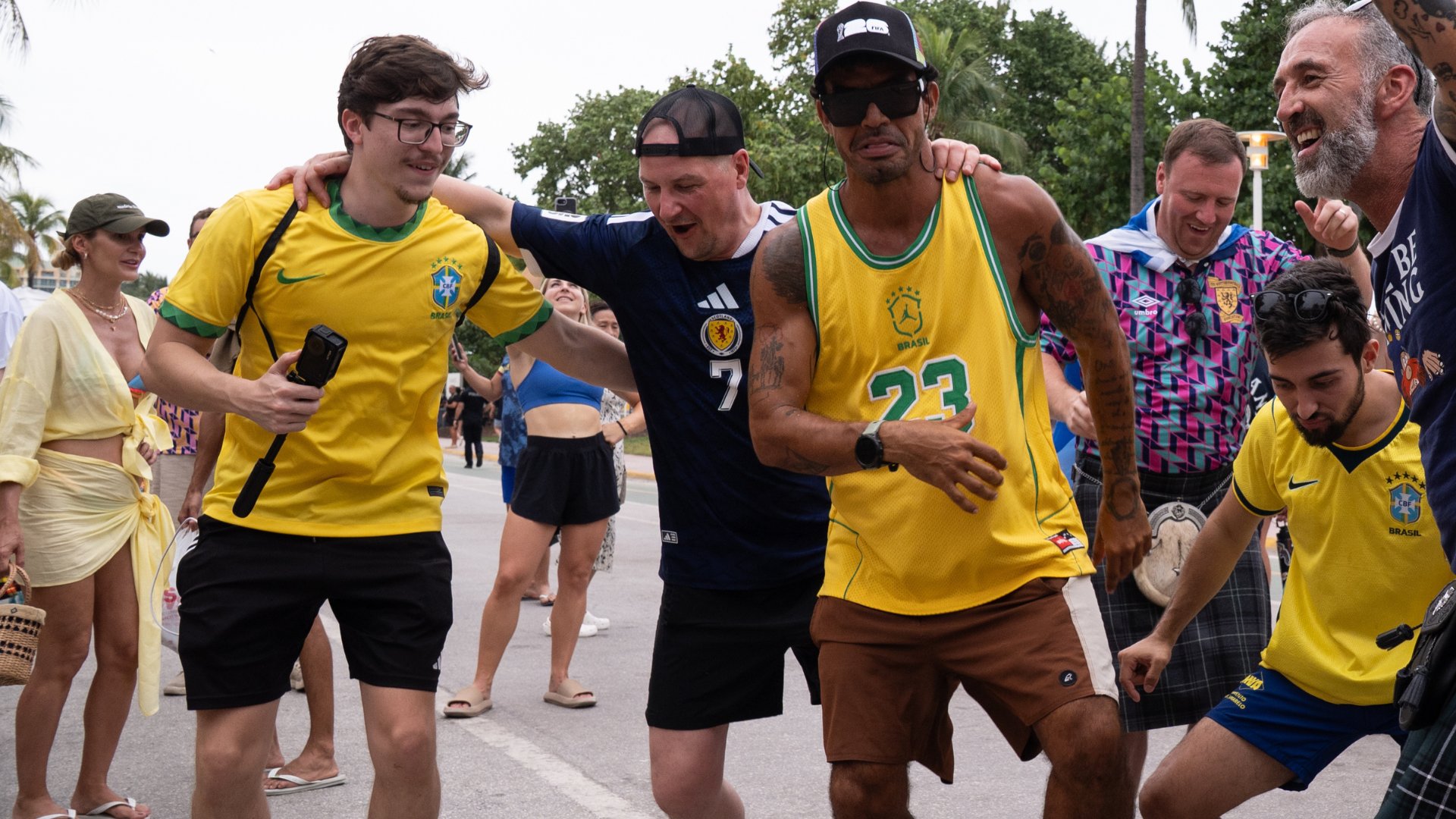 Fans in yellow Brazil jerseys and a Scotland jersey walk arm-in-arm down a street, smiling together in a celebratory crowd.