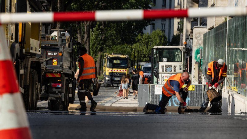 Workers in orange high-visibility vests clean a street alongside construction vehicles and barriers.