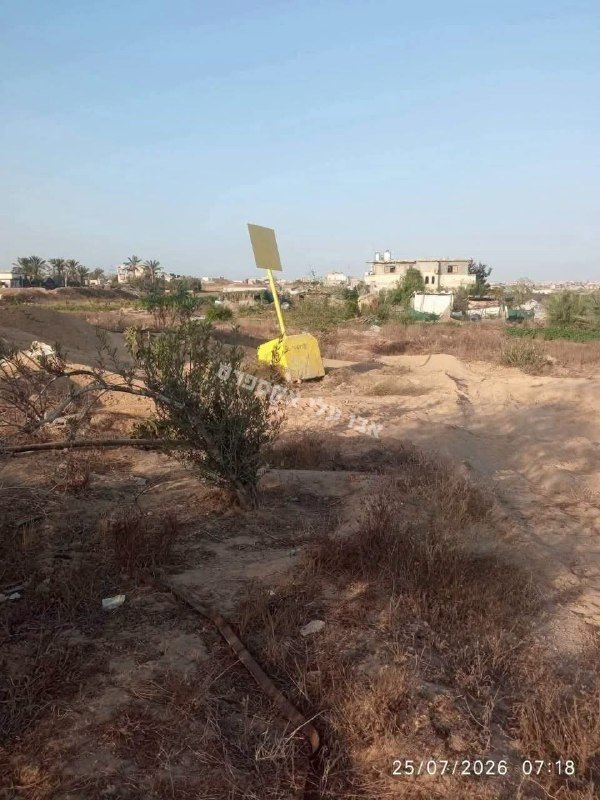 A yellow marker post stands in a dry, sandy field with sparse vegetation, foregrounding a distant view of low-rise buildings under a clear sky.