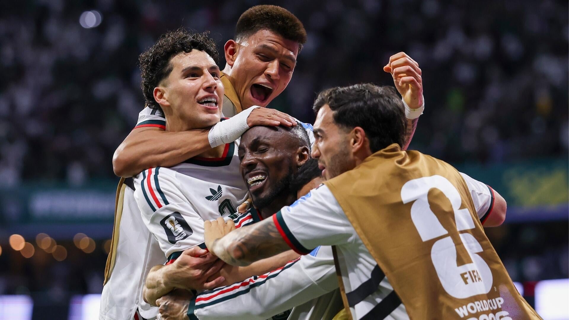Four soccer players in white jerseys with red and green trim embrace and shout in celebration, one wearing a gold "FIFA World Cup 2025" bib.