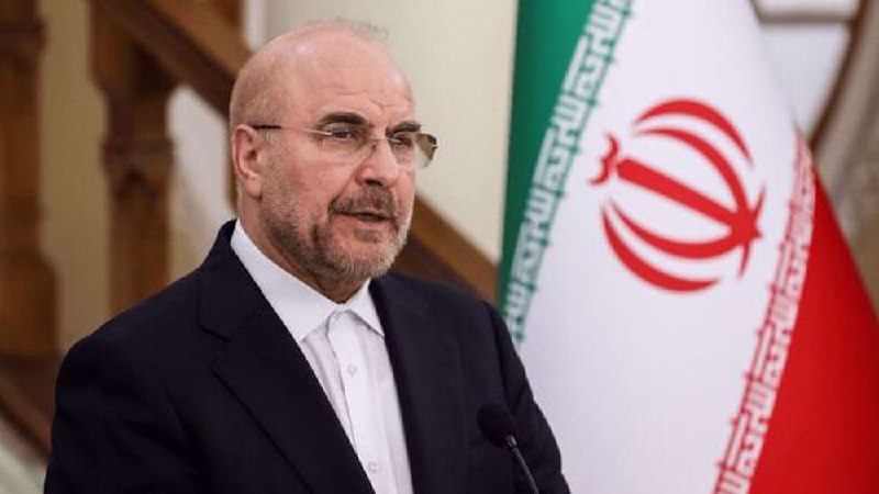 A bald, bearded man wearing glasses and a dark scarf stands in a crowd of men in suits.