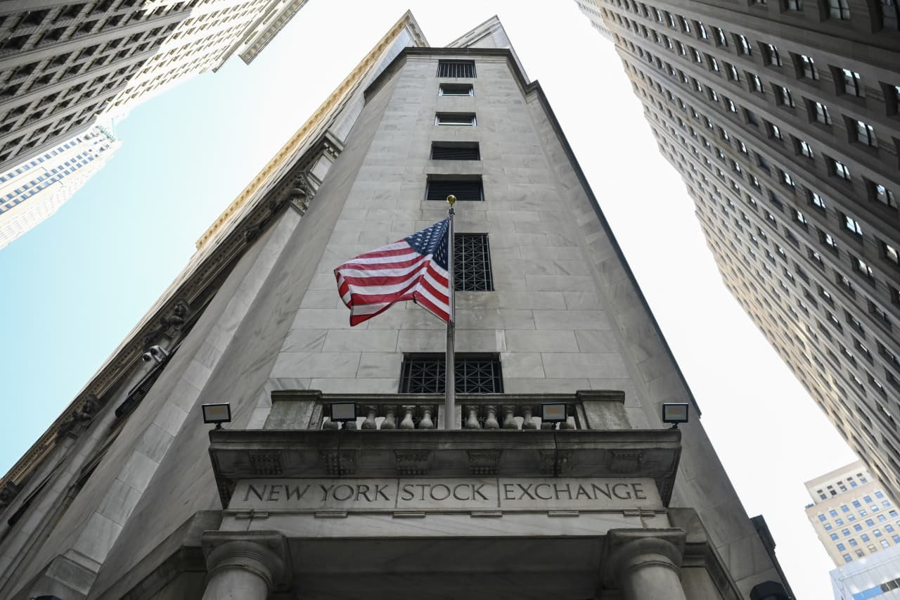 A low-angle view shows the facade of a stone building inscribed "NEW YORK STOCK EXCHANGE," with an American flag flying from a central pole, flanked by tall skyscrapers.
