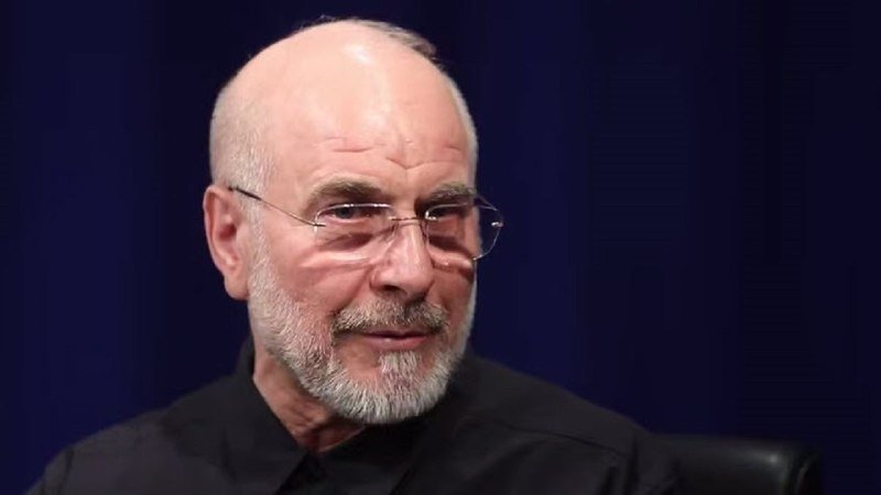 A bald, bearded man wearing rimless glasses and a black shirt speaks against a dark blue background.