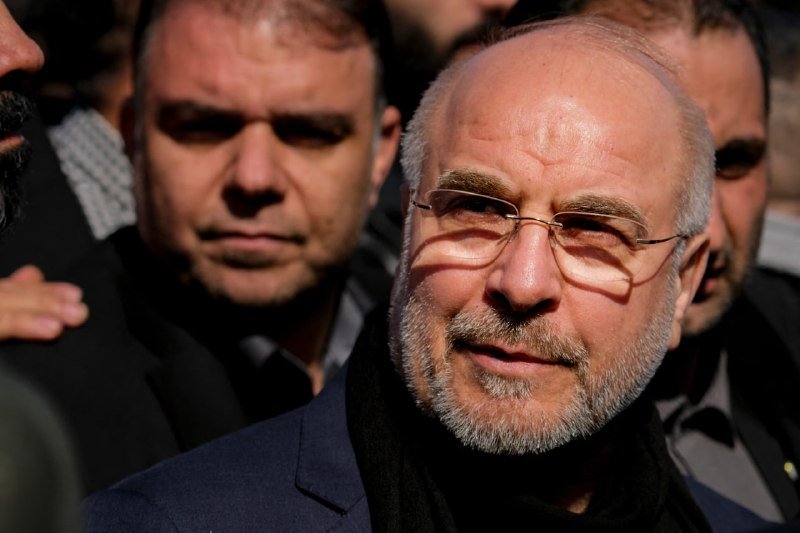 A balding, bearded man wearing rimless glasses and a dark coat stands in a crowd of men outdoors, with sunlight casting a lens-flare across his face.