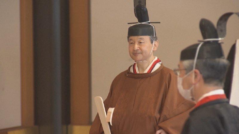 Man wearing traditional Japanese ceremonial attire with tall black headpiece holds a folded white fan indoors.