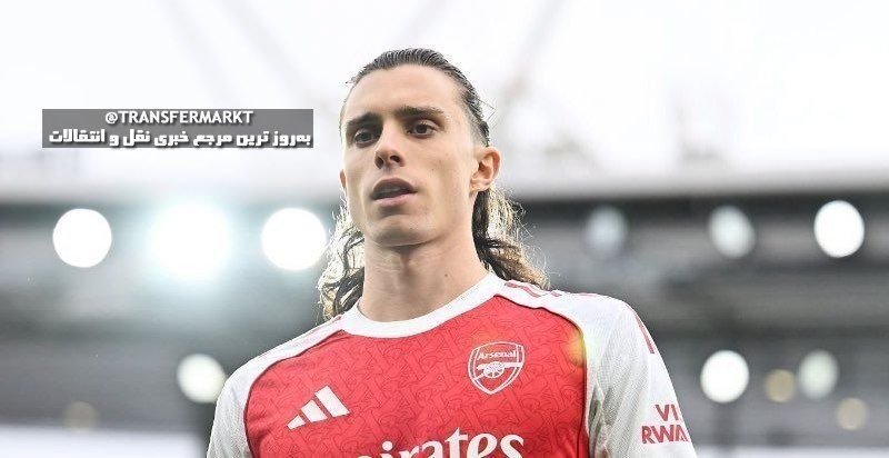 A long-haired male soccer player wearing a red Arsenal jersey stands in a stadium setting.