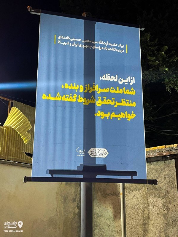 A nighttime outdoor billboard displays a Persian-language poster with yellow and white text, featuring a blurred face graphic and a portrait outline, mounted on a pole beside a wall.