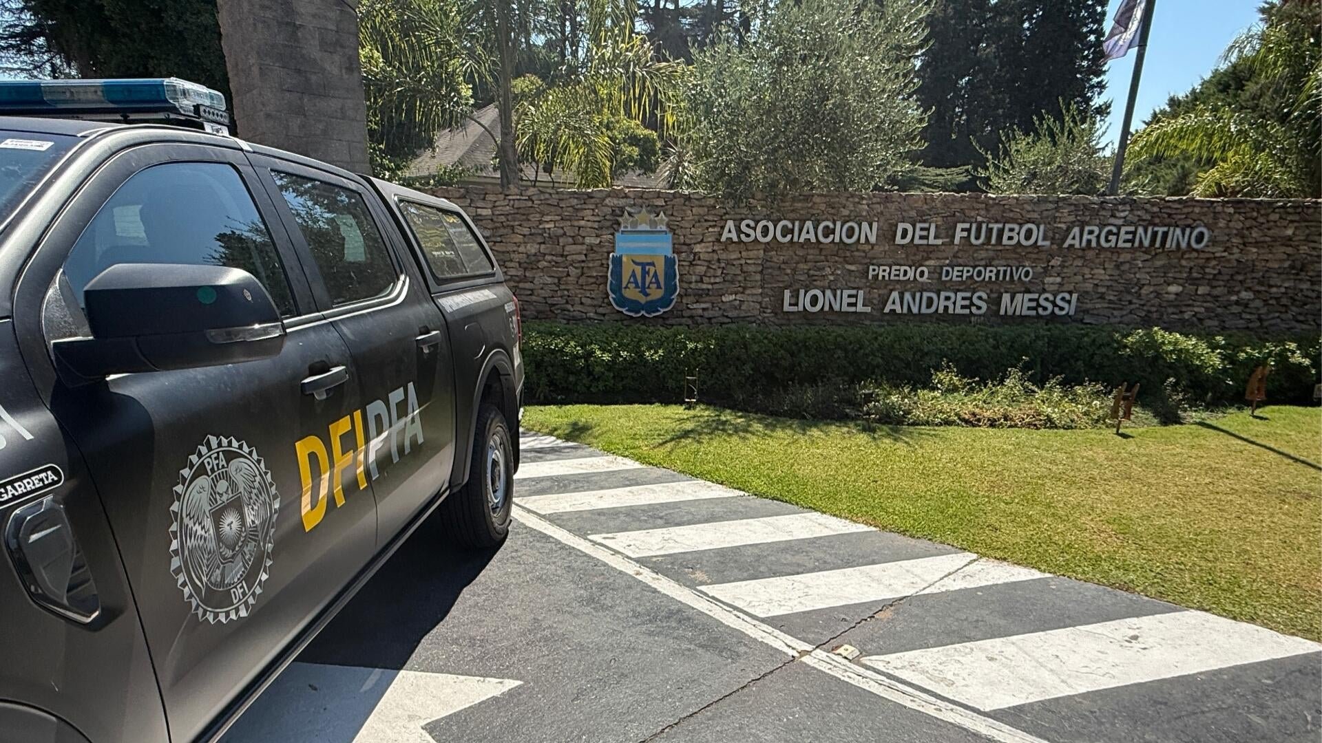 A black police vehicle labeled "PFA DFI PFA" is parked near a stone wall sign reading "ASOCIACION DEL FUTBOL ARGENTINO PREDIO DEPORTIVO LIONEL ANDRES MESSI."