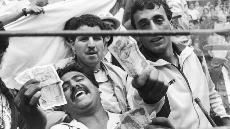 Black-and-white photo of three cheering men in a crowd holding up paper currency, with a microphone visible in the foreground.