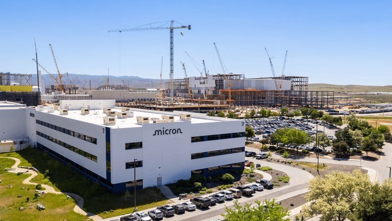 A white building labeled "micron" stands beside a large construction site with multiple cranes, surrounded by parking lots and mountains in the background.