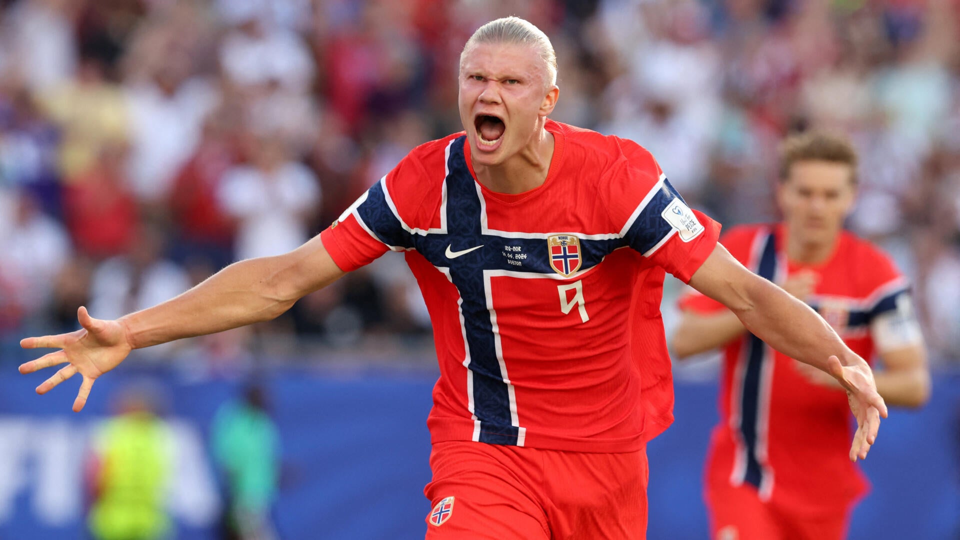 A soccer player wearing a blue and green Brazil jersey with "BRASIL" written on it warms up near a stadium sideline, with photographers and spectators in yellow visible behind him.