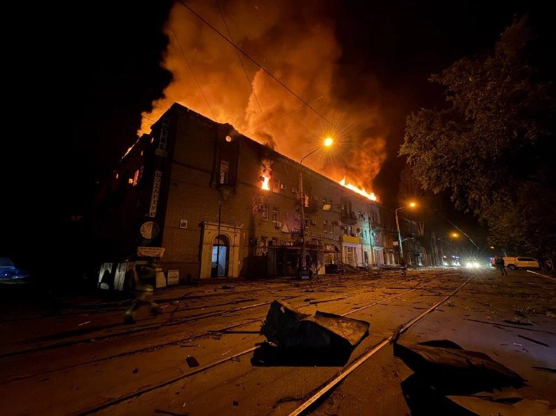 Flames and thick smoke engulf a multi-story building along a nighttime street, with debris scattered across the pavement and tram tracks.