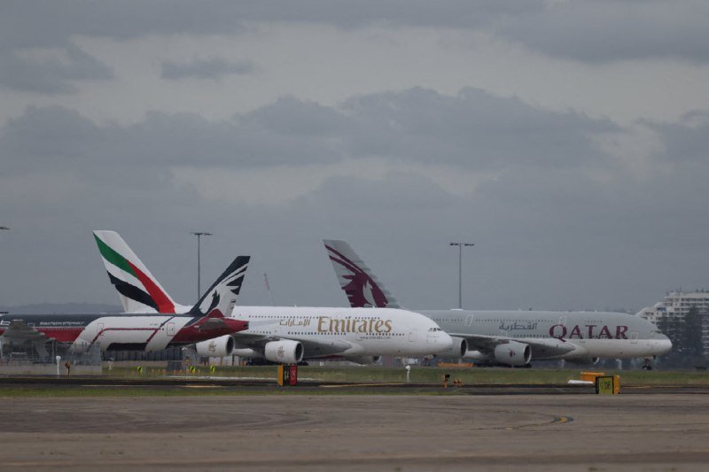 Two large commercial passenger jets, labeled "Emirates" and "Qatar," sit on an airport taxiway under a cloudy sky.