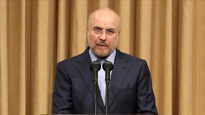 A bald, bearded man in a dark suit and light blue shirt speaks at a podium with two microphones, set against beige curtains.