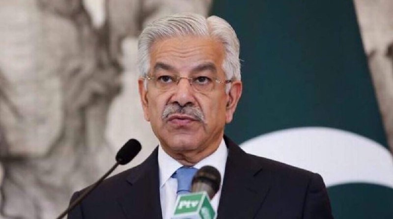 A gray-haired man with a mustache and glasses speaks at a podium with microphones, with a green and white flag visible behind him.