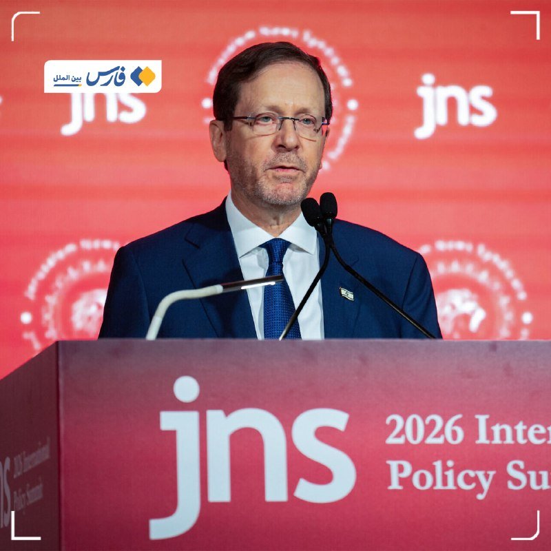 A bespectacled man in a dark suit and blue tie speaks at a podium labeled "jns" and "2026 Inter... Policy Su..." against a red "jns" branded backdrop.
