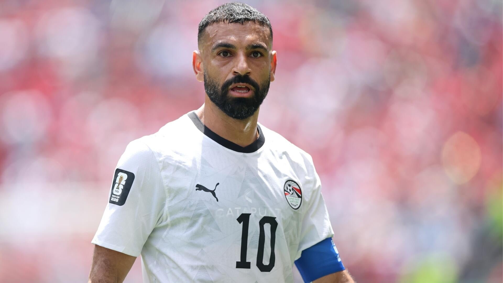 A bearded soccer player wearing a red jersey with the number 10 leaps into the air in celebration while a teammate in a red number 22 jersey runs behind him on a stadium field.