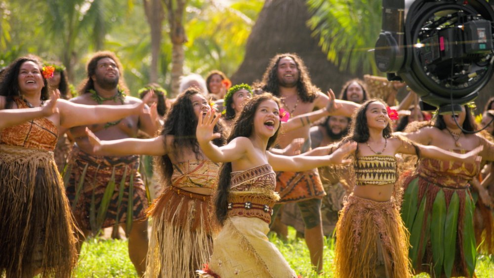 A group in traditional grass skirts and floral tops dances outdoors among palm trees while a large film camera records them on set.