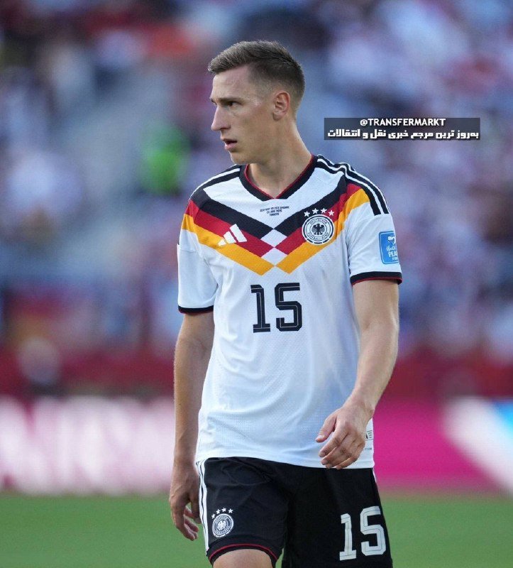 A male soccer player wearing a white Germany national team jersey with the number 15 stands on a pitch with a blurred stadium crowd behind him.