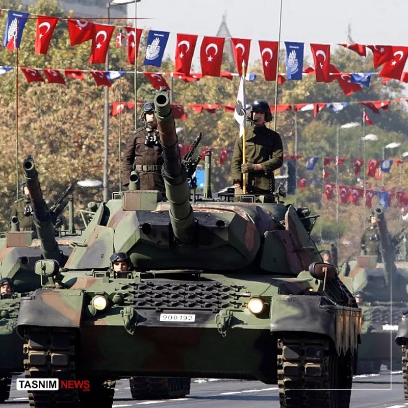 Two soldiers stand atop a camouflaged military tank marked "900192" during a parade, with Turkish and other national flags strung overhead.