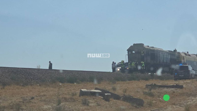 Emergency responders in high-visibility vests gather near a derailed train on a dry, grassy hillside beside railroad tracks, with a police vehicle parked nearby.