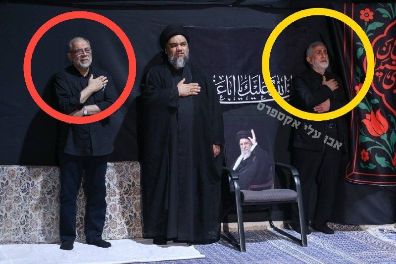Two men stand with hands over their hearts while a cleric in black robes and turban stands between them on a stage with Arabic calligraphy and a framed portrait.