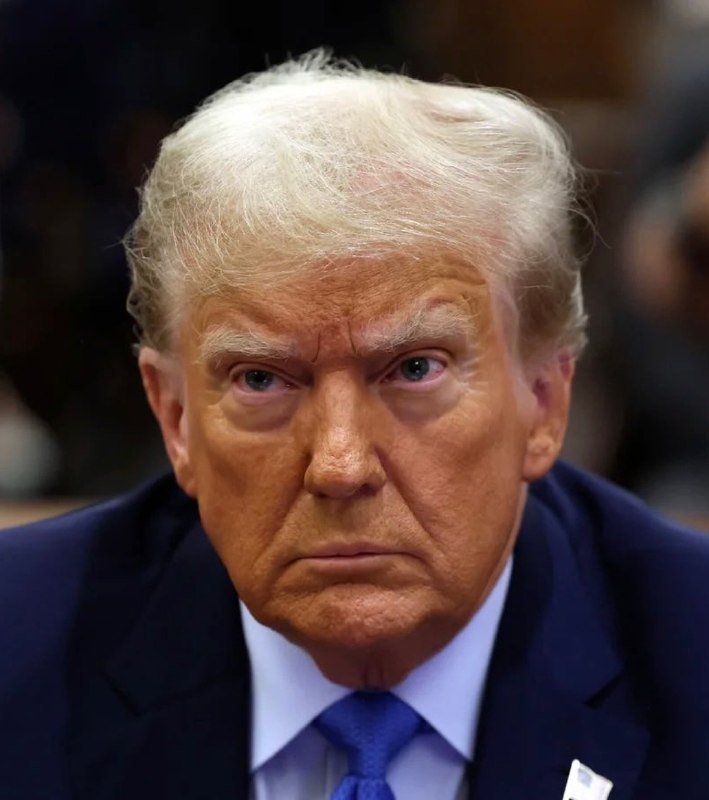 A man with blonde hair wearing a dark blue suit and blue tie looks forward with a serious expression.