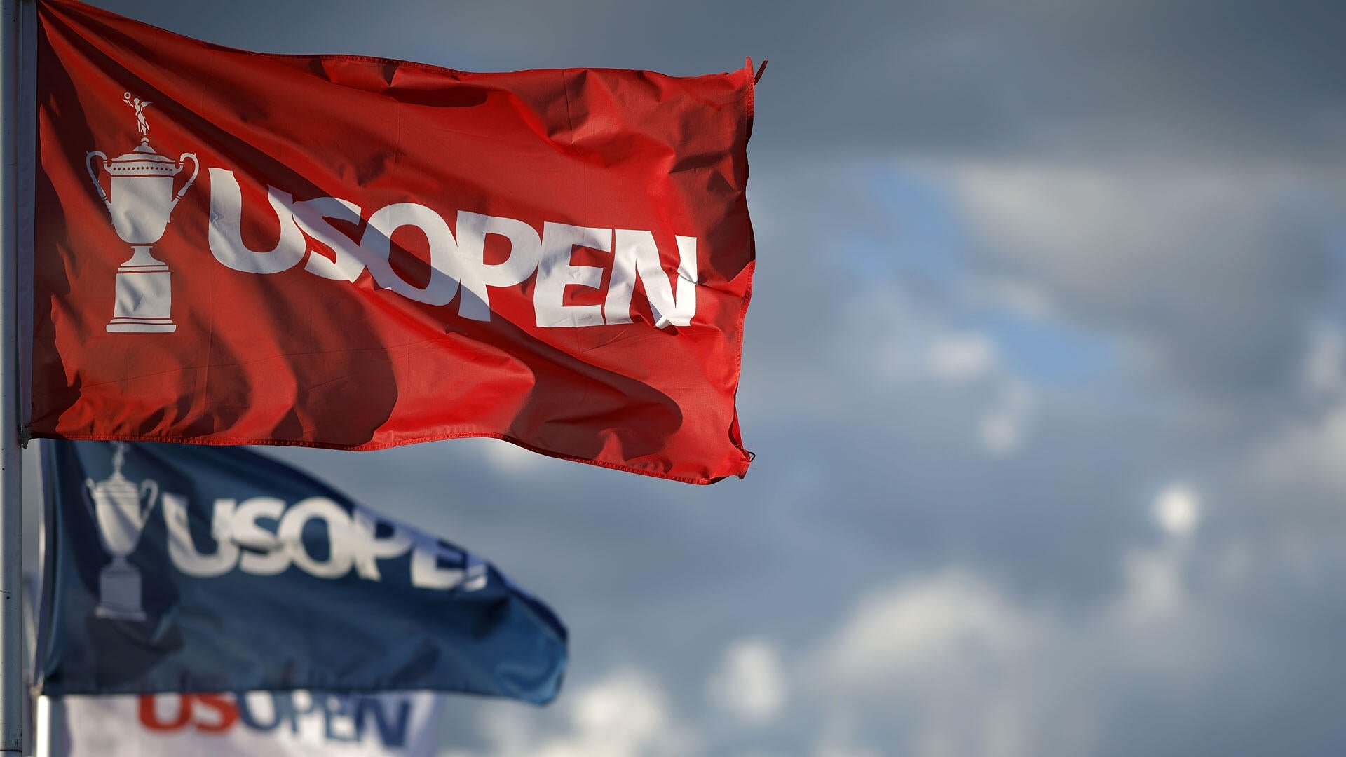 Red, blue, and white flags displaying the "US OPEN" logo and trophy emblem wave on a flagpole against a partly cloudy sky.