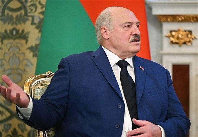 A bald man with a mustache in a blue suit gestures while speaking, seated before a red-and-green flag in an ornate room.