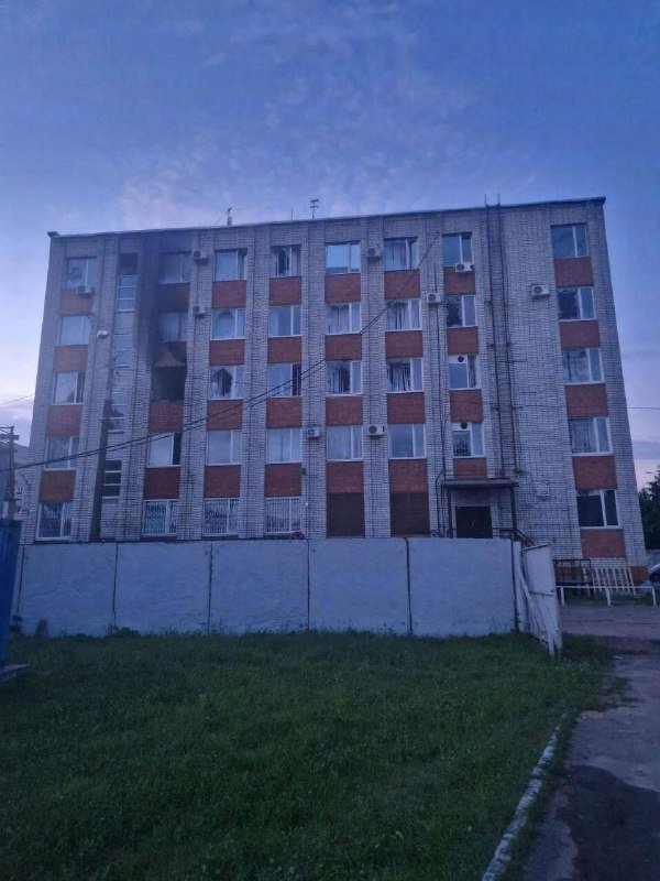 A five-story brick apartment building with multiple windows and air conditioning units stands behind a concrete wall, surrounded by overgrown grass under a cloudy blue sky.