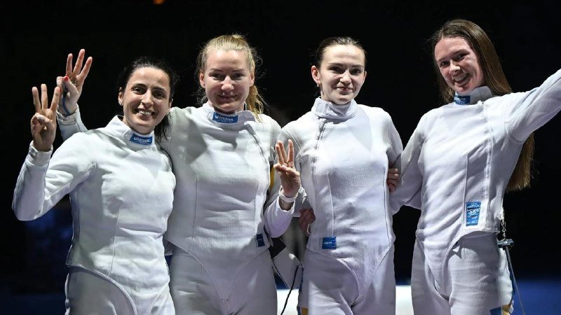 Four women in white fencing jackets smile and pose together, with two holding up fingers to indicate a score.