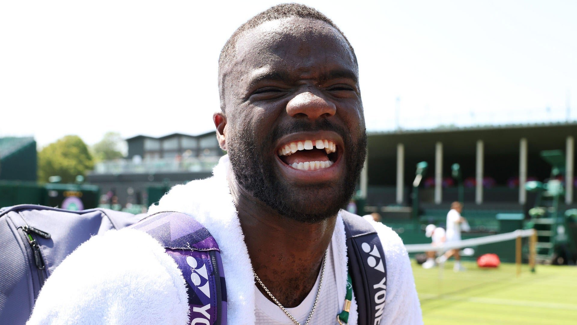 A man with a beard, carrying a Yonex tennis bag and wearing a white towel, smiles broadly on a grass tennis court.