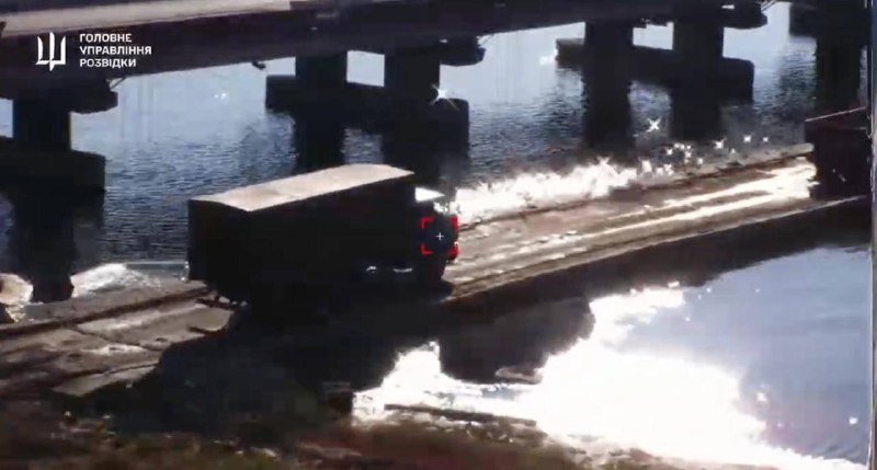 A dark military truck drives across a damaged bridge spanning a river, with white Ukrainian text and a trident logo in the upper left corner.