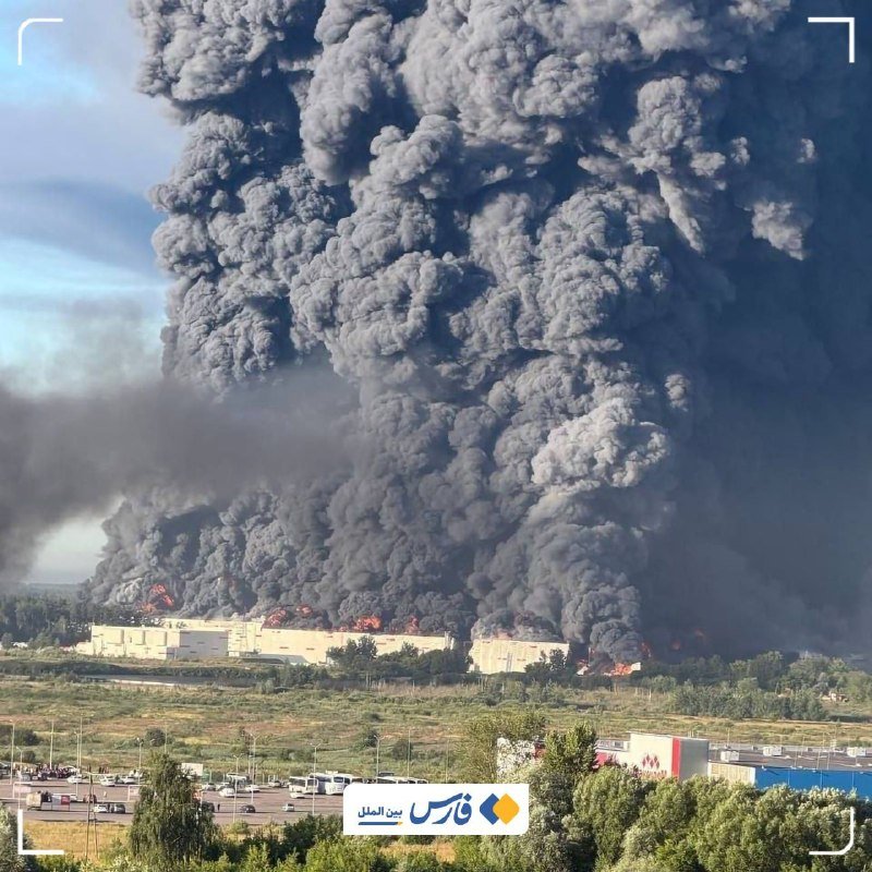 Massive gray smoke billows from a large burning industrial building, with visible flames along its roof, overlooking a nearby road and smaller commercial structures.