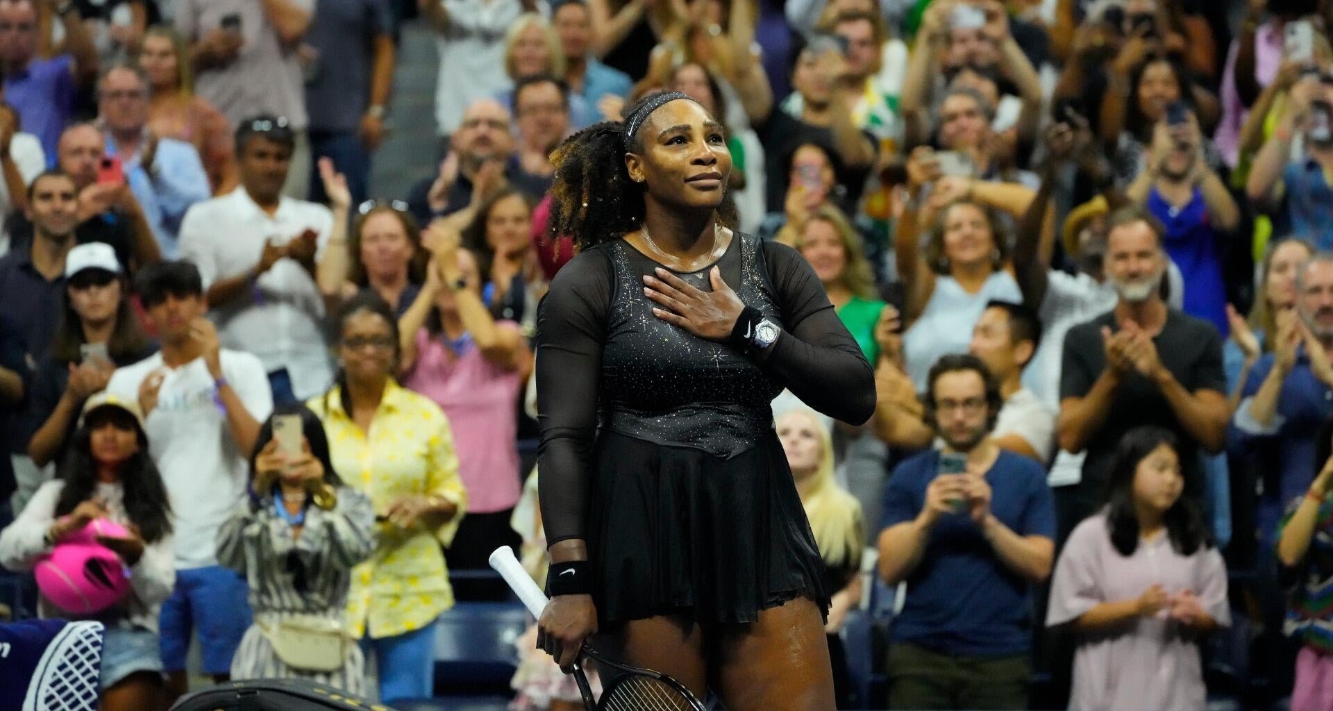 A female tennis player in a black outfit stands on court with her hand over her heart, looking upward as the crowd behind her applauds and takes photos.