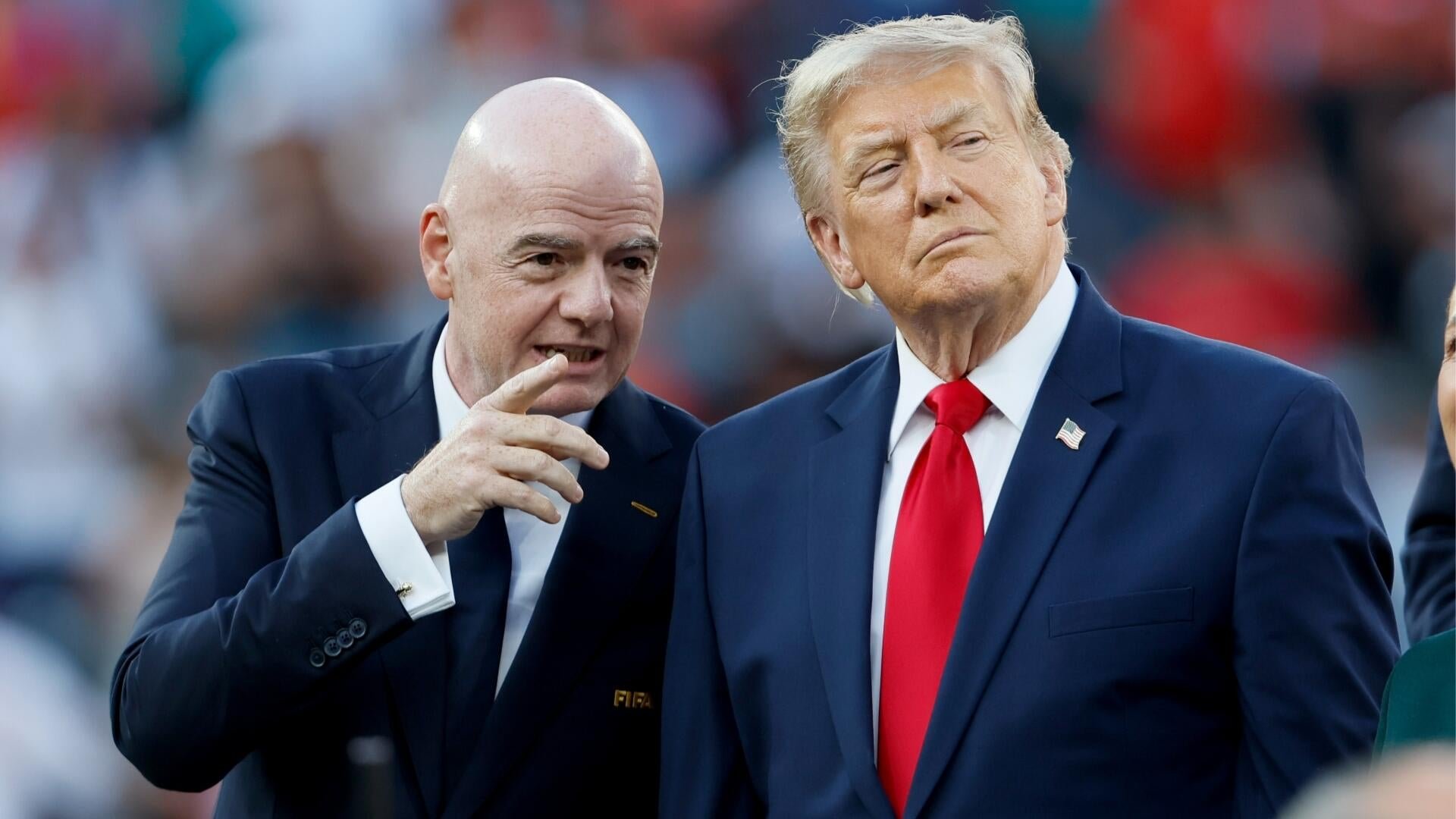 Two men in suits stand side by side, one gesturing with his hand while speaking, the other wearing a red tie and an American flag pin, against a blurred crowd background.