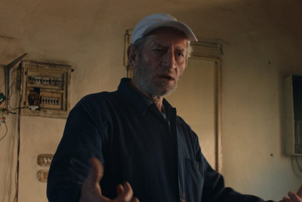 A bearded man wearing a white cap and dark jacket gestures with his hands inside a room with exposed electrical panels on the wall.