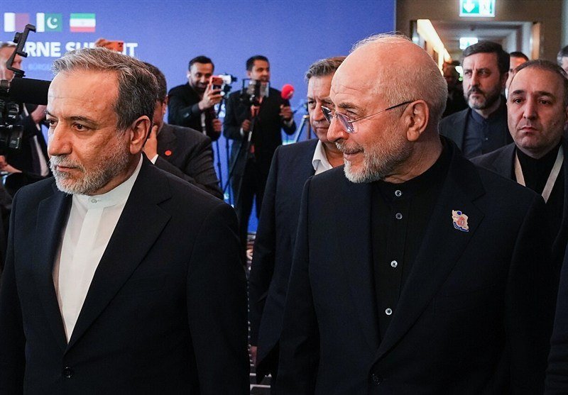 Two men in dark suits and ties stand back-to-back against a light-colored building wall, with one looking downward and the other gazing forward.