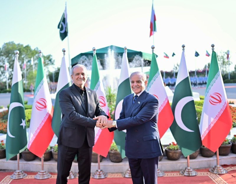 Three small desk flags of Iran, Pakistan, and the United States stand on a wooden surface against a blurred background.