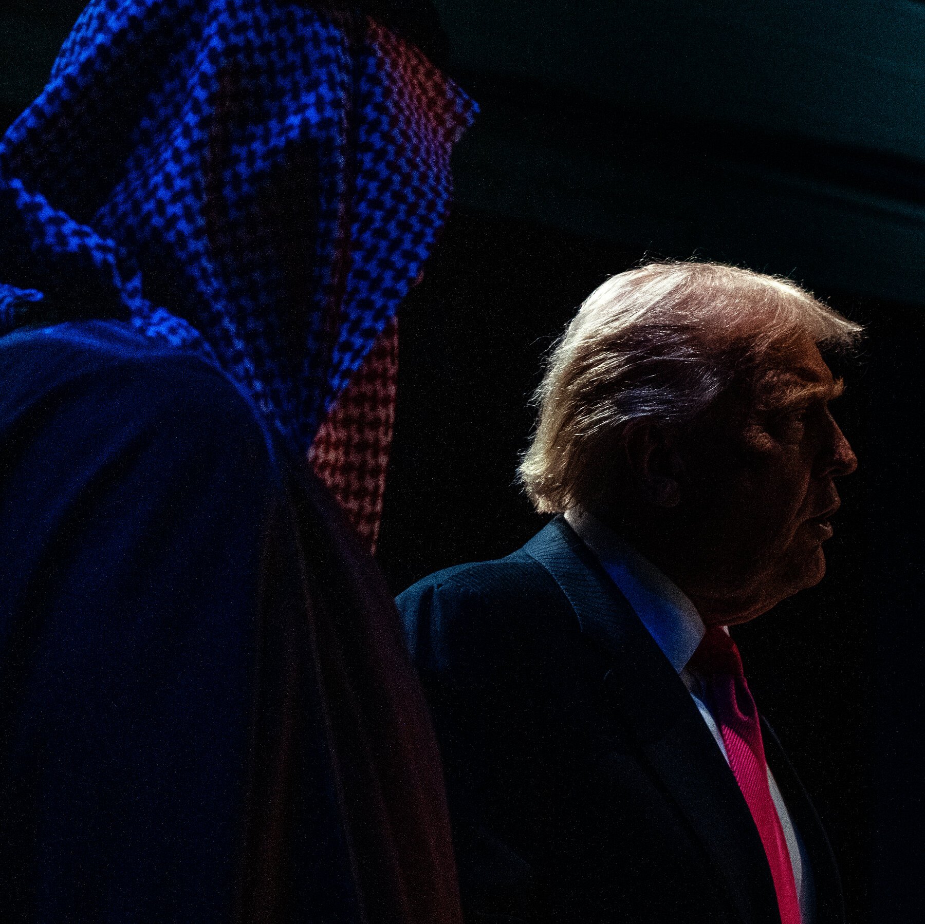A man with blond hair in a dark suit and red tie stands in profile, silhouetted against a dark background with a blue-lit keffiyeh displayed to his left.