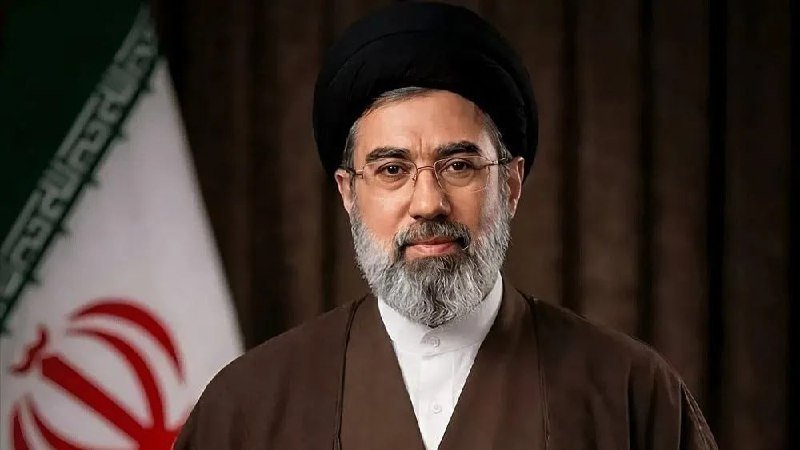 A bearded man wearing glasses, a black turban, and brown clerical robes stands in front of an Iranian flag.