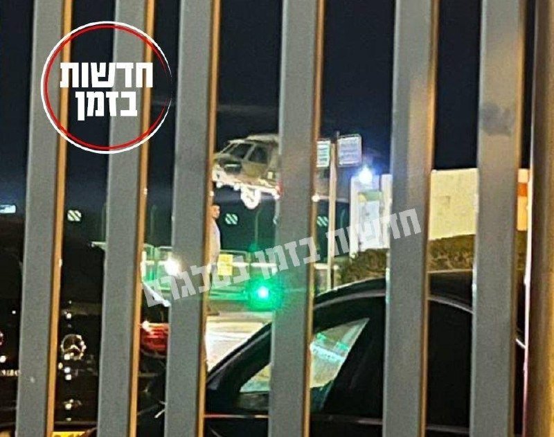A tow truck carries a white vehicle at night, viewed through a metal fence, with Hebrew text watermarked across the image.